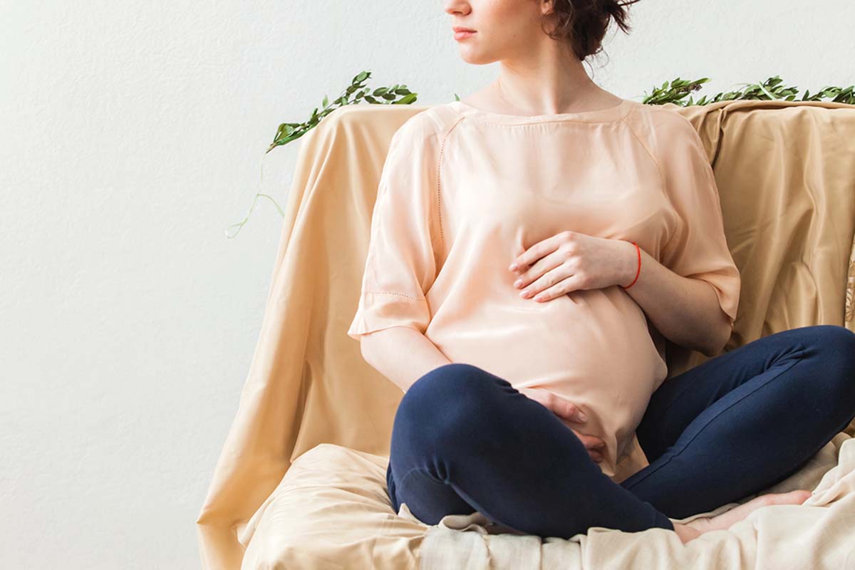 A seated pregnant woman in a peach blouse and blue pants rests her hand on her belly while looking off to the side. The couch is draped with a light fabric. Greenery accents the background.