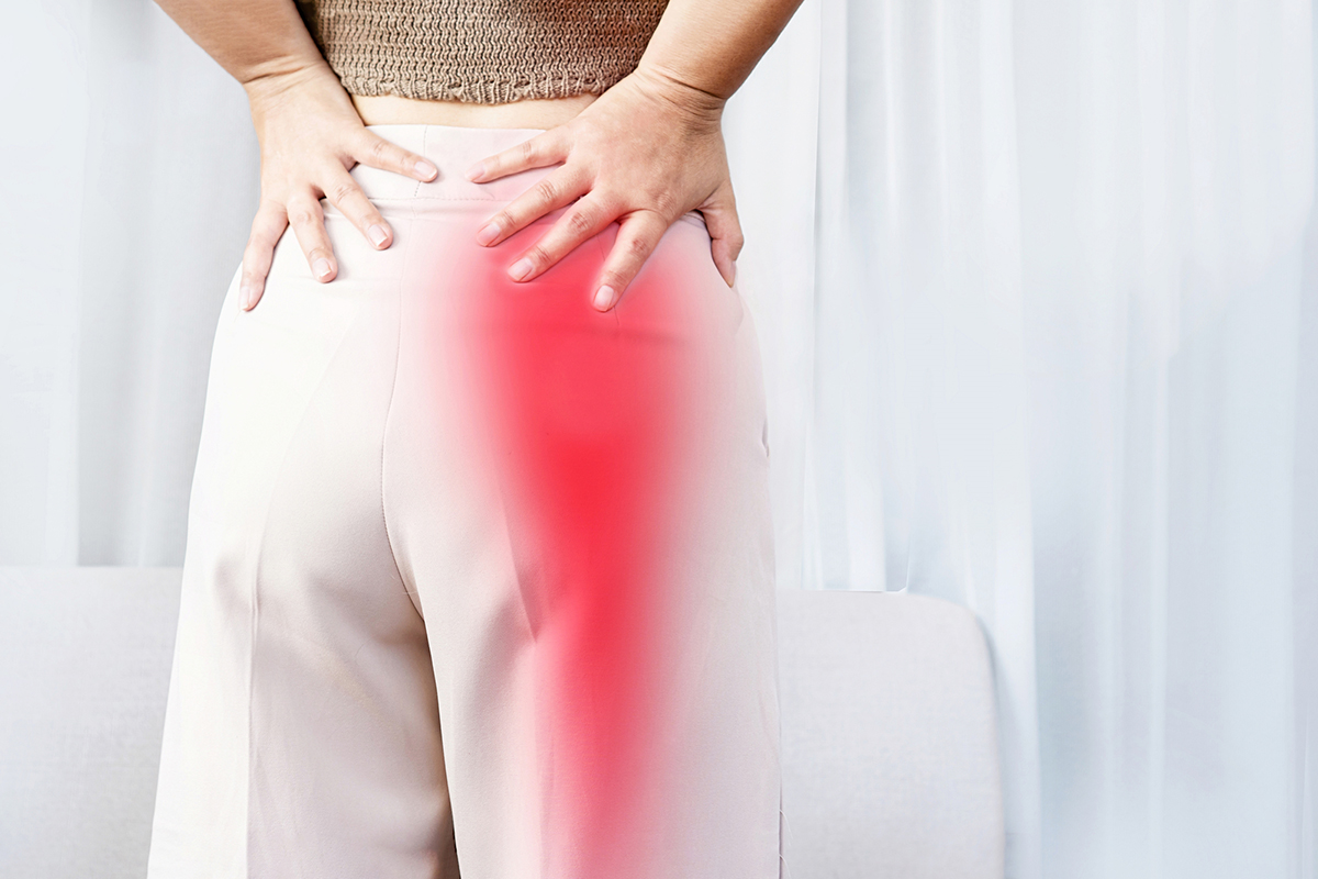 A person is holding their lower back with both hands, with a red highlighted area indicating pain extending from the lower back to the upper leg.