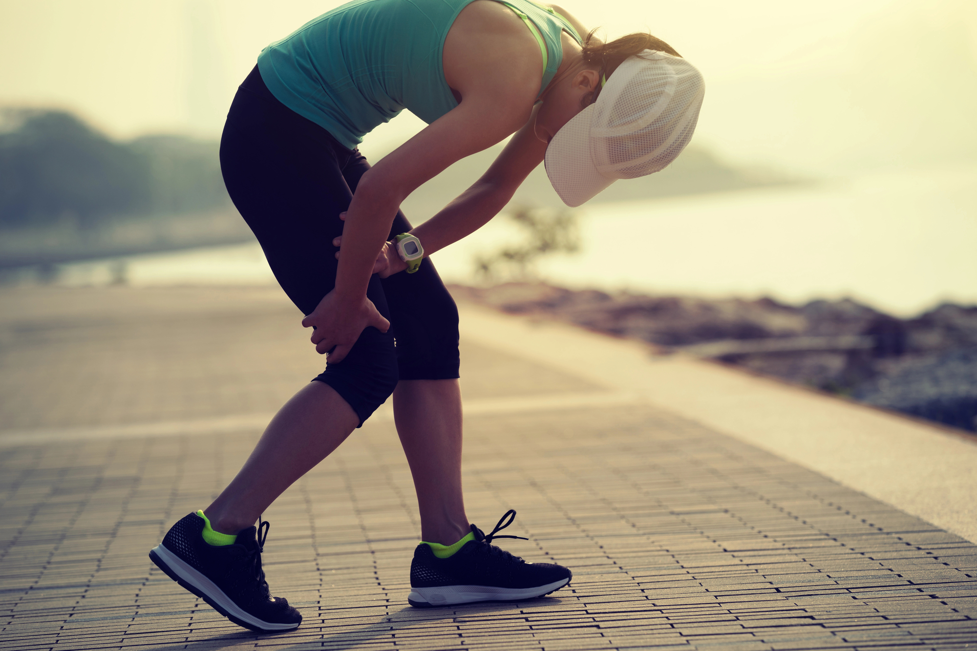 A person wearing a tank top, leggings, and a cap bends over holding their knees while standing on a paved outdoor path near water.
