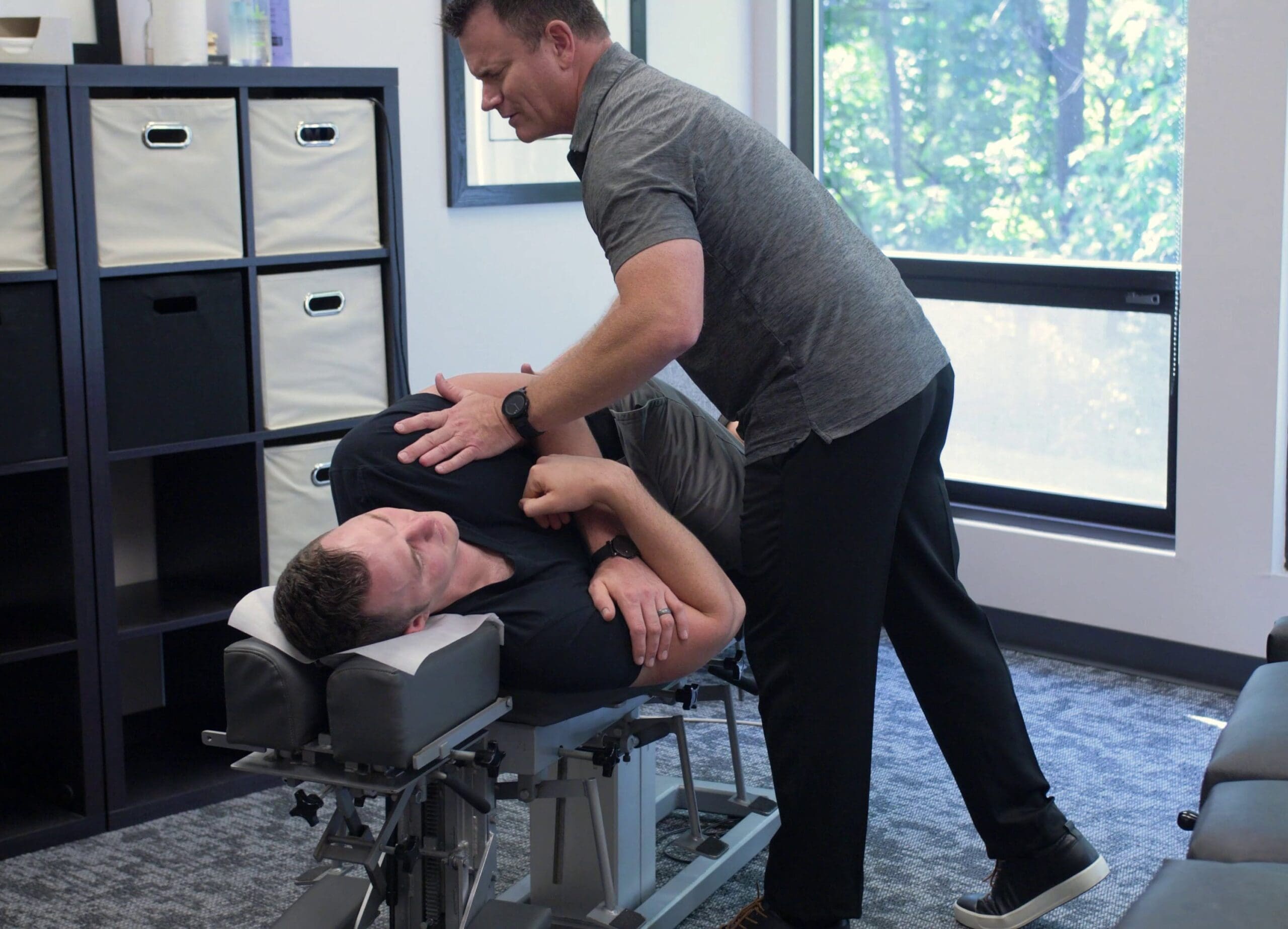 A physical therapist assists a male patient lying on a treatment table, performing a shoulder manipulation exercise in a clinic.